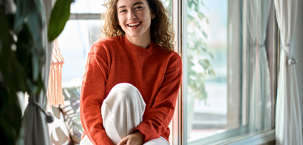 Jeune femme aux cheveux bouclés, pull orange, souriant devant une fenêtre ensoleillée à Muret en Haute-Garonne 31